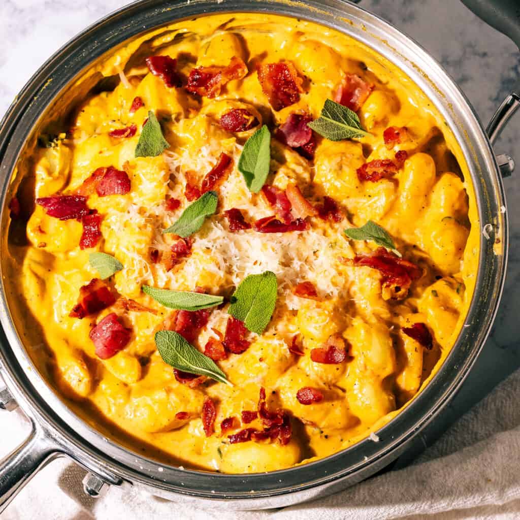 Large skillet full of creamy pumpkin gnocchi topped with bacon and sage leaves.