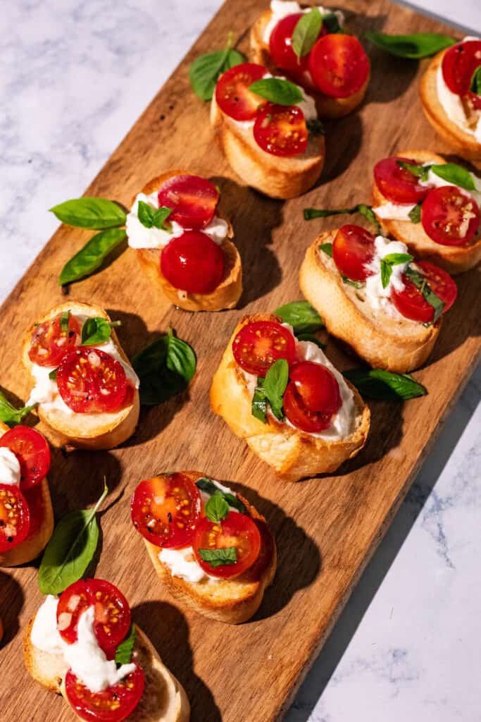 Bruschetta toasts topped with tomatoes, burrata, and basil on a long wooden plank.