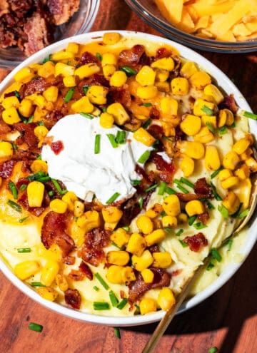 Bowl of mashed potatoes topped with sour cream, corn, bacon, chives, and cheese with a gold spoon.