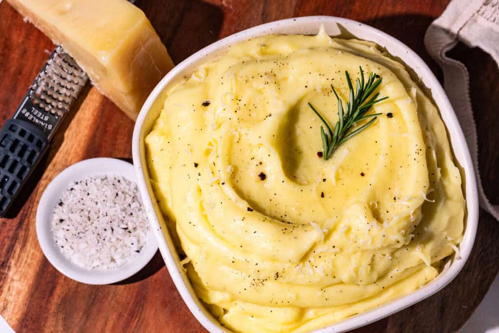 A bowl of truffle mashed potatoes garnished with rosemary and a side of truffle salt and parmesan cheese.