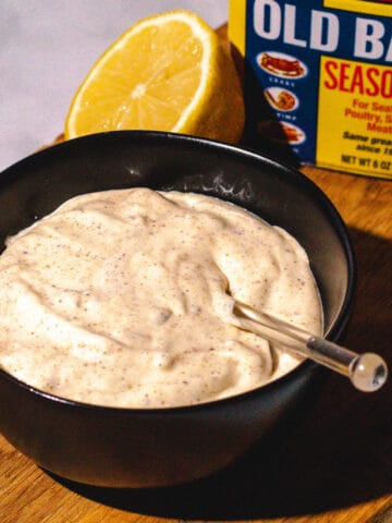 Tartar sauce in a black bowl with a container of Old Bay in the background.